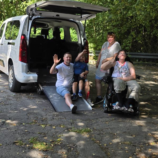 Eine Familie winkt vor einem geöffneten Kofferraum mit einer Rollstuhlrampe